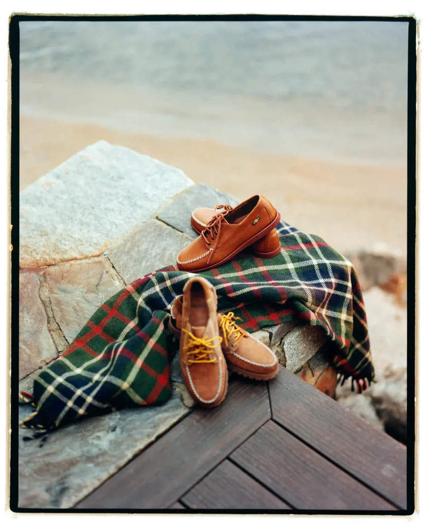 En primer plano se ven un chal de hilo decorado a cuadros y dos pares de zapatos tipo náutico colocados de forma cuidadosa, al borde de una tarima de madera que remata junto a un suelo de piedra. De fondo se aprecia la zona de la orilla del mar en una playa de arena, con el agua en calma.